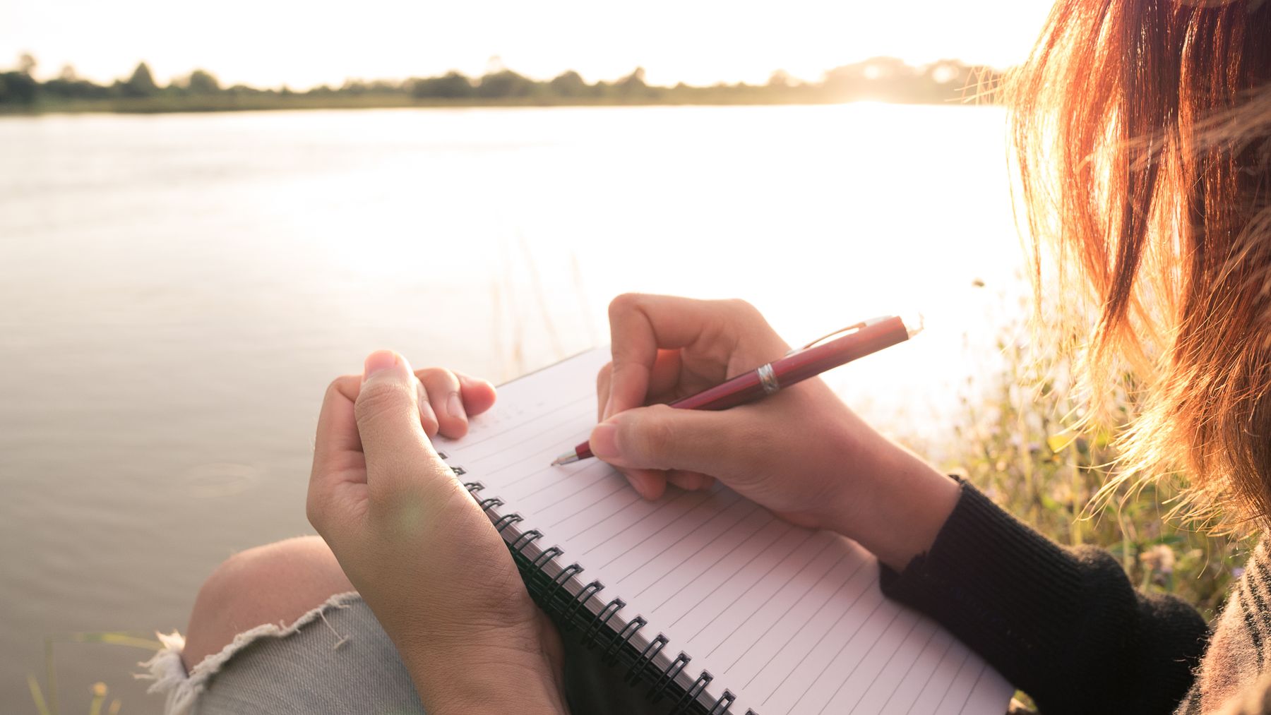A hand writing in a notebook beside a lake