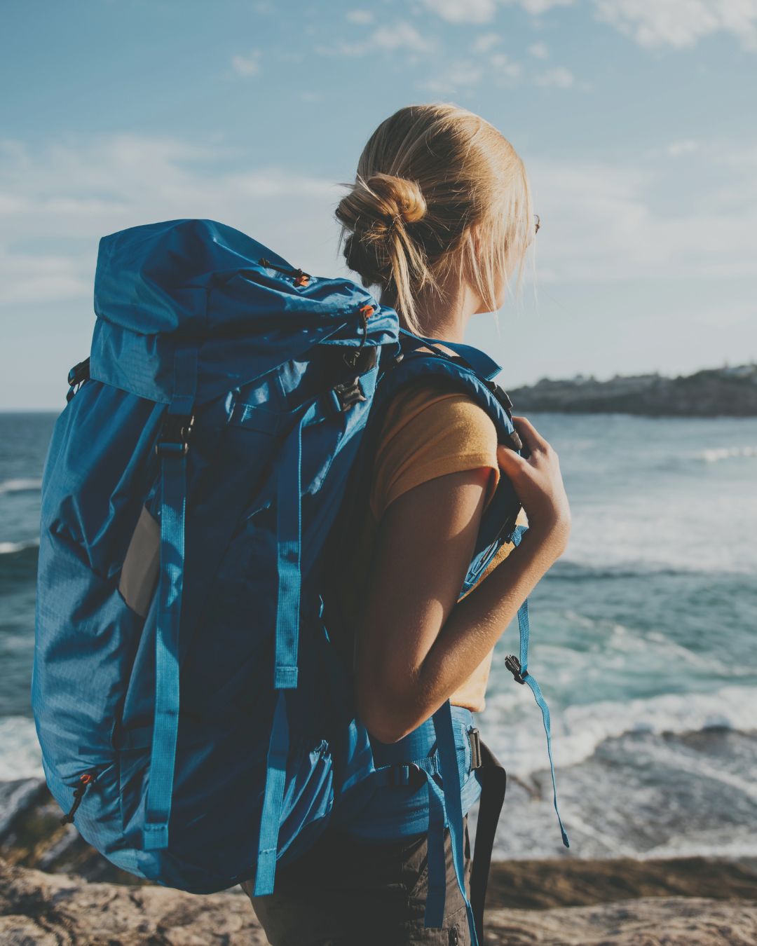 A woman carrying a large blue backpack, looking out to sea